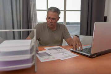 Mature man sits with laptop, paperwork, addressing electricity bill dispute, he is anger,depicting responsibility and problem-solving in modern life,he has grey hair.