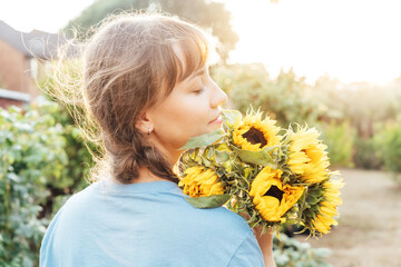 Dreaming young female farmer woman with closed eyes holding and sniffing a sunflowers bouquet on...