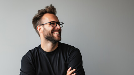 A young man with a beard and glasses is wearing a black t-shirt, smiling and looking away from the camera. The background is a light gray color