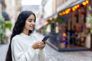 Close-up photo of a young Indian woman standing on a city street smiling and using a mobile phone