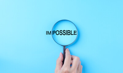 Hand holding magnifying glass focuses on possible from the word impossible on a light blue...