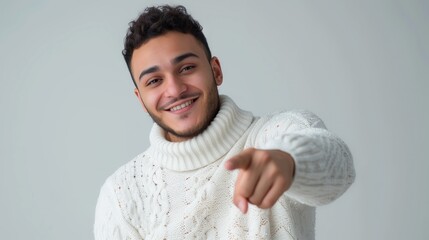 Confident young man with dark hair and a well-groomed beard, wearing a white turtleneck sweater, smiling and pointing at the camera on grey background - obrazy, fototapety, plakaty