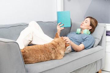 Attractive girl in headphones reading book and her cute dog lying close to her. Audiobook and home...