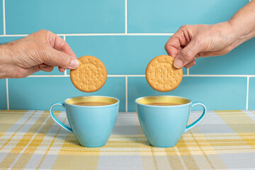 Cups of tea in attractive blue and yellow cups with hands dunking rich tea biscuits. Cups isolated against a blue tiled background and sitting on yellow check. Mugs of hot drink with a sweet treat.  - obrazy, fototapety, plakaty