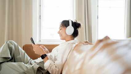 Relaxed woman enjoying music on headphones while using smartphone, comfortable at home.