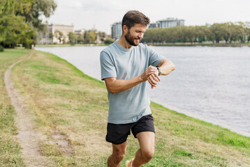 Male jogger checking his watch during a workout by the river, surrounded by greenery.