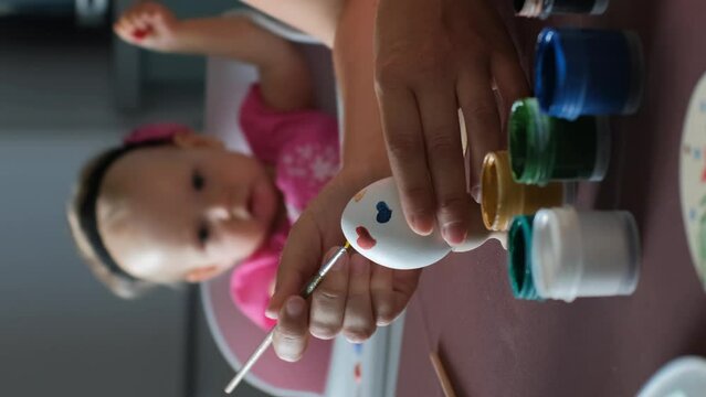 Vertical video. Baby playing while mother painting Easter egg in the kitchen, selective focus