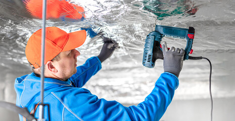 The builder is fastening the vapor barrier under the ceiling thermal insulation using an electric...