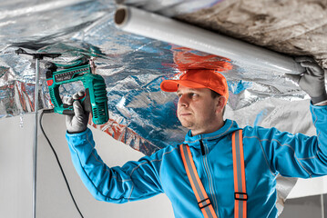 The builder is fastening the vapor barrier under the ceiling thermal insulation using an electric...