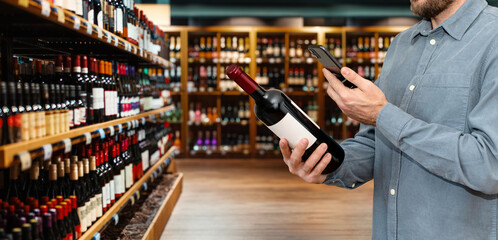 Man in casual clothes customer in liquor store scans e-label on wine bottle using his smartphone. - obrazy, fototapety, plakaty