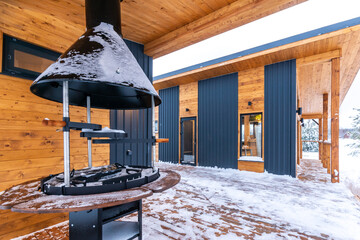 An outdoor terrace with a barbecue oven and a wooden chalet house covered with snow on a winter day.