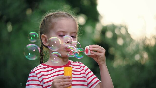 Joyful little girl with thin braids blows soap bubbles in surrounding of blurry garden. Colorful soap bubbles flying around carelessly under gaze of little girl. Girl spends free time blowing bubbles