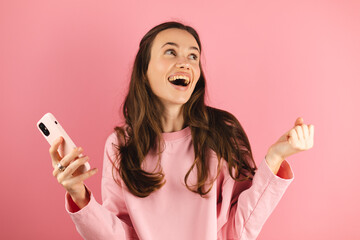 Amazed brunette woman look surprised wow hold using mobile cell phone on pink background. Girl look...