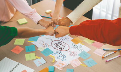 Young happy creative startup group join circle fist bump hands together surrounded by marketing strategy mind map and colorful sticky notes at meeting room. Unity and teamwork concept. Variegated.
