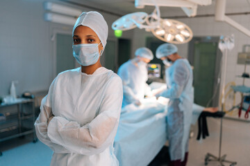Portrait of confident African-American female doctor standing posing with crossed arms looking at camera in operating theater. Diverse team of surgeons performing operation on background.