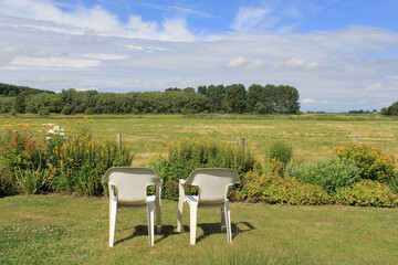 two white chairs at a lawn in the flower garden with a beautiful view at a green meadow and trees and blue sky in the background