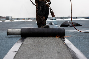 professional roofer at work securing rolled tar paper with a blowtorch on a flat roof installation...