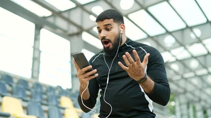 Disappointed athlete checking results of training while looking at smartphone standing in urban city stadium. Upset male in tracksuit unhappy with negative workout results or bad news on mobile phone
