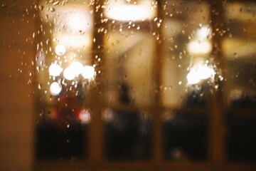 Abstraktes Motiv mit Regentropfenmuster vor Glasfassade mit braunen Rahmen von Restaurant mit Gästen und weißen Lichtern bei Regen am Abend im Winter