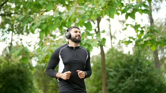 Adult bearded male runner in wireless headphones jogging outside in urban city park. Handsome man in sports suit likes to run in the fresh air while listening to his favorite music. Healthy lifestyle