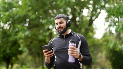 Adult bearded smiling sportsman walking with headphones and mobile phone enjoying listening to music outdoors on urban city park. Happy joyful male athlete in black sportswear relaxing after training
