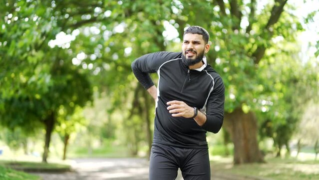 Adult bearded runner suffering from back pain, sports injury, muscle spasm in urban city park. A handsome sportsman in sportswear having back flank ache and problem after exercise outside in nature