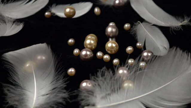 White and pink pearl beads fall on the black velvet with swan feathers laying on it, slow motion 