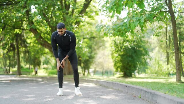 Handsome male runner experiences muscle pain and discomfort during a jog in the city park. He is massaging and stretching to alleviate the trauma injury he may have sustained while jogging outdoors