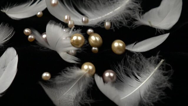 White and pink pearl beads fall on the black velvet with swan feathers laying on it, slow motion 