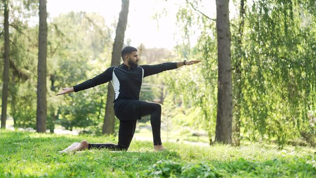Adult sportsman does gymnastics while standing barefoot on a mat in an urban city park. Sporty male practices yoga outside. Fit athlete in a tracksuit performs exercises in the forest after work