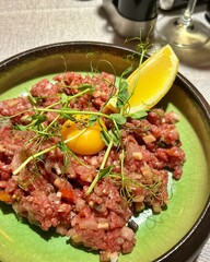 plate with raw meat tartare, tartare with finely chopped pickled cucumbers, egg yolk, tartare, lemon wedge, pea sprouts