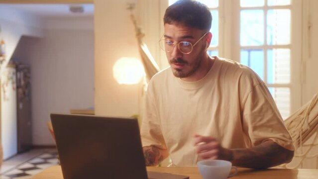 Young tattooed man in glasses and wireless earphones waving, showing thumbs up and speaking on video call on laptop in living room with warm light, having e-meeting or spending weekends at home
