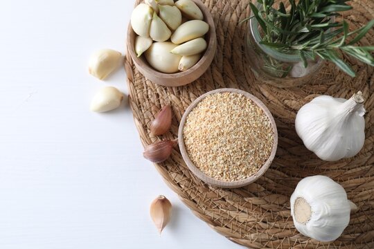 Dehydrated garlic granules in bowl, rosemary and fresh cloves on white table, flat lay. Space for text