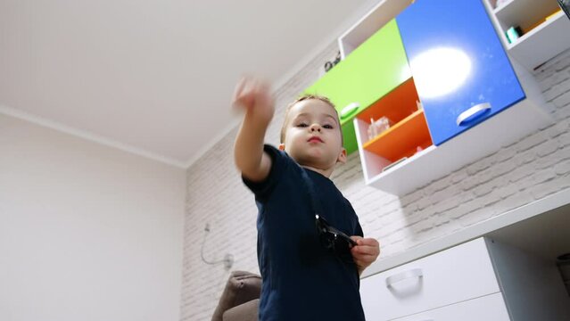 Beautiful baby boy holding sunglasses stands in the room. Little kid counting fingers. Low angle view.