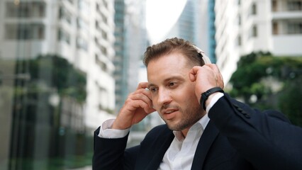 Businessman put on headphone and listen relaxing music while sitting at green city with blurred background . Skilled manager project manager enjoy to listen while dance to song in lively mood. Urbane.