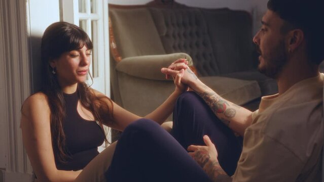 Young pretty woman holding hand of boyfriend, sharing her thoughts and smiling while sitting on the floor in a doorway in the evening at home