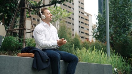 Businessman wear headphone to listen relaxed music while take off suit and move to music at green city. Happy project manager with headset dancing movement with lively song while hold phone. Urbane.