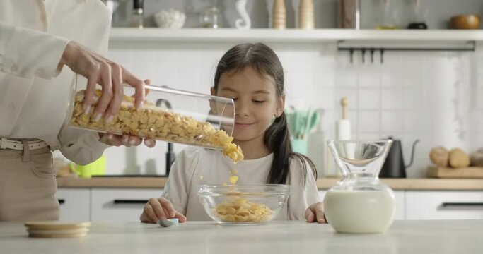 Mother pours corn flakes into kid's bowl at breakfast. Woman hands prepare cereal for smiling girl in the kitchen at home. Cereal corn flakes