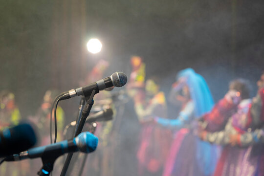 A foreground microphone takes center stage amidst a sea of blurred colors. In the background, vibrantly dressed individuals dance on a stage, capturing the effervescent energy of a carnival ensemble