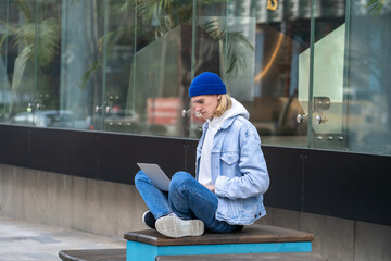 Pensive hipster sitting in street in yoga stance with laptop on knees, working as freelancer,...