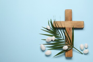 Wooden cross, painted Easter eggs and palm leaf on light blue background, flat lay. Space for text