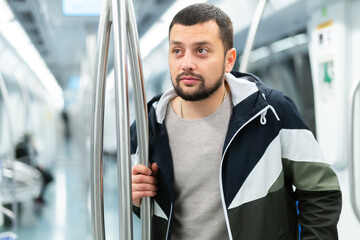 Portrait of thoughtful young bearded man holding on handrails in subway car during daily commute to work ..