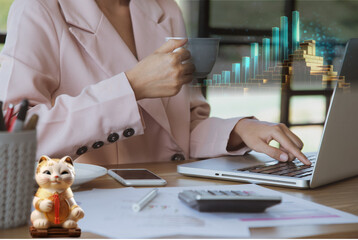 Mujer de negocios trabajando en su laptop tomando su taza de café, sobre el escritorio vemos su móvil, su calculador y su gato de la suerte. Un gráfico de barras muestras sus ganancias.