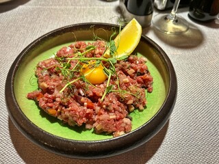 plate with raw meat tartare, tartare with finely chopped pickled cucumbers, egg yolk, tartare, lemon wedge, pea sprouts