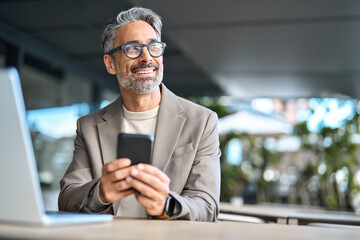 Happy middle aged business man executive using mobile phone sitting outside office. Stylish older busy businessman investor wearing glasses holding smartphone looking away at copy space with cellphone - obrazy, fototapety, plakaty