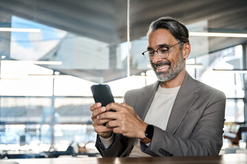 Smiling mature business man executive sitting at desk using smartphone. Happy busy middle aged...