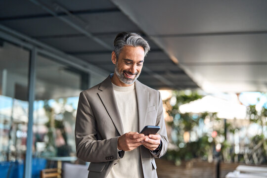 Happy mature older male entrepreneur standing outdoors looking at smartphone. Smiling middle aged business man, rich professional businessman holding cellphone using mobile cell phone digital tech. - Powered by Adobe