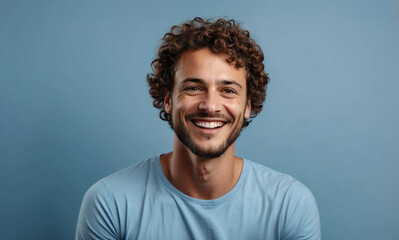 Portrait of handsome 30 year old brown curly haired guy with wide smile and light blue t-shirt, looks at camera with happy expression