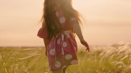Joyful little girl in summer dress runs in field under rays of sunset. Happy child spends vacations...