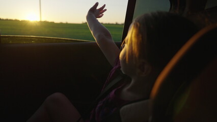 Little girl sits in car, looks out window and waves her hand. Happy family enjoy car travel outdoors, together vacation. Daughter dreams of adventure, looks out open car window. Childrens journey